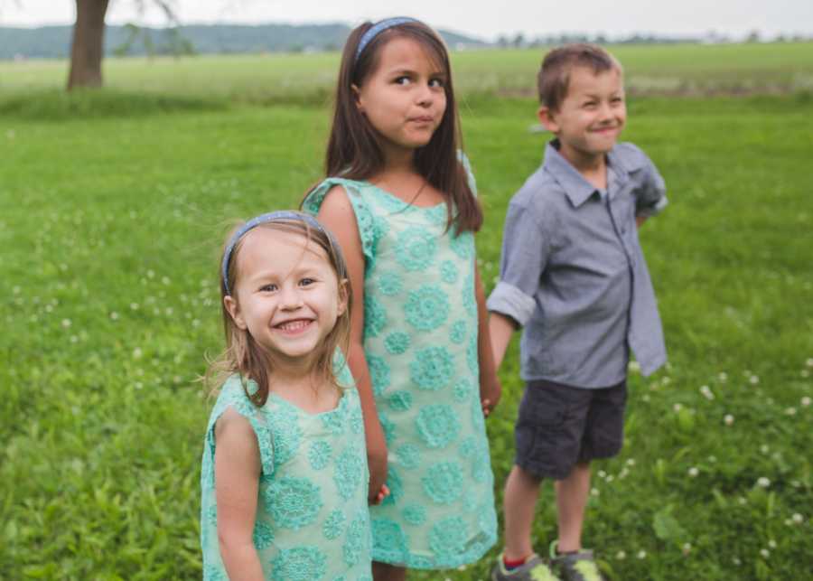 Three young siblings hold hands while standing outside whose mother has sworn in front of them