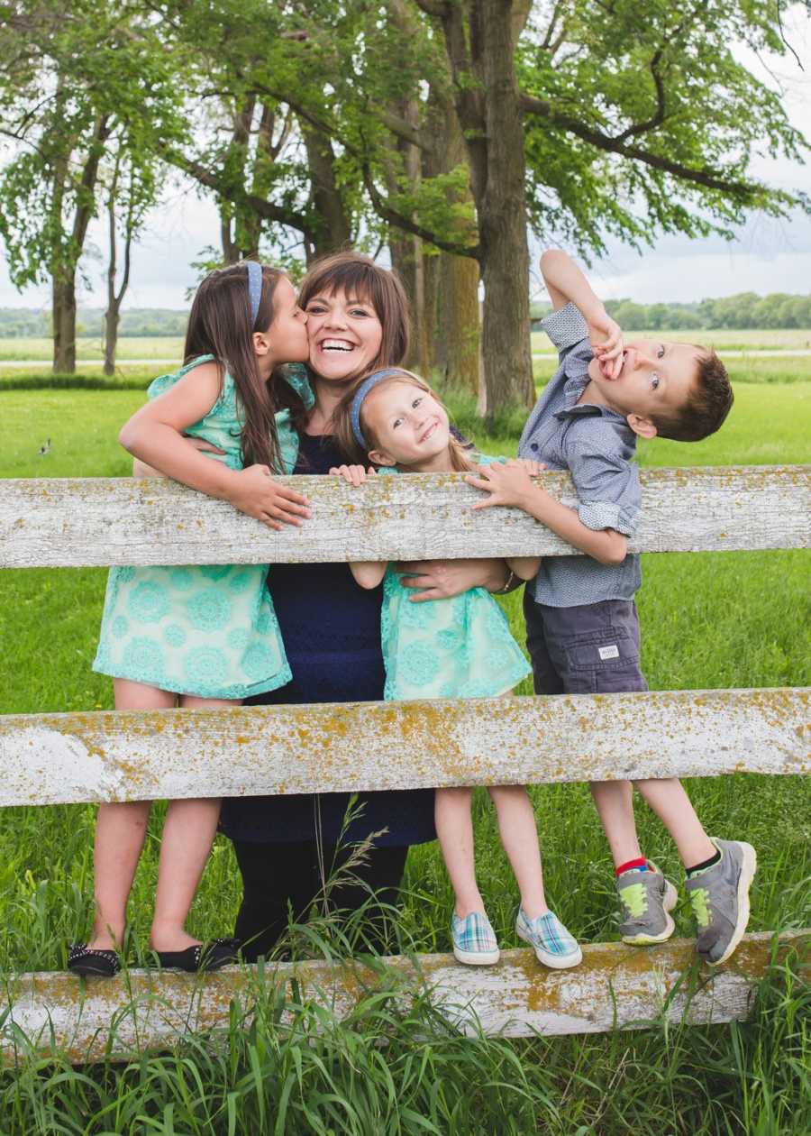 Mother smiles as her oldest child kisses her on the cheek while her two younger kids hold onto wooden fence 