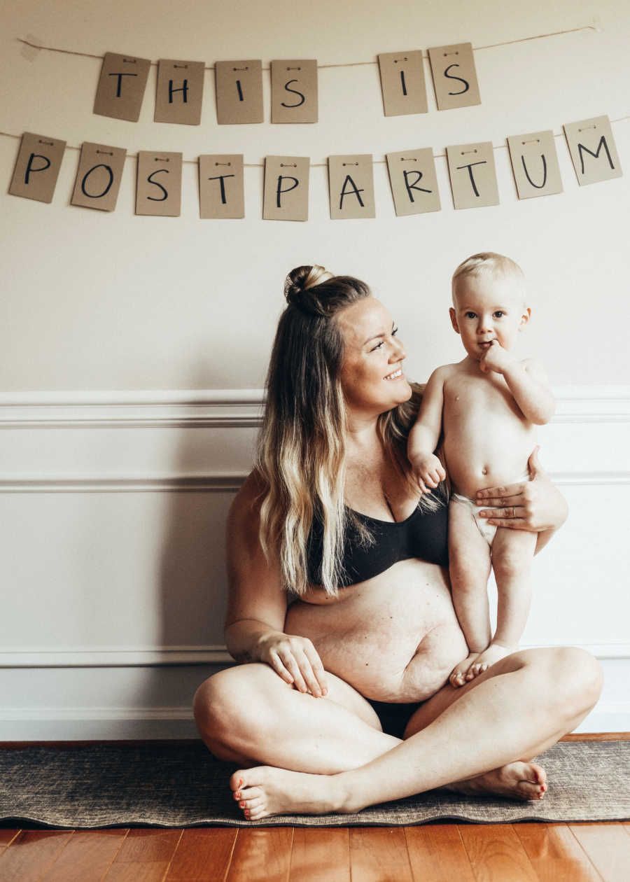 Woman sits in bra and underwear on floor with son standing on her legs below banner saying, "This is postpartum"