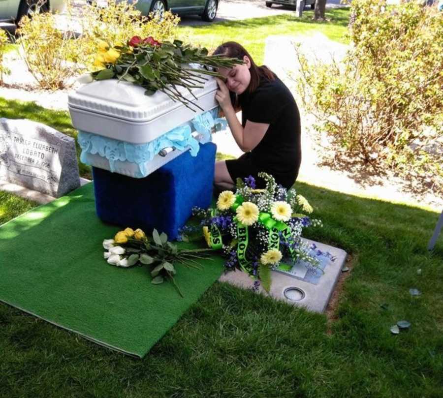 Mother kneels on ground resting her head on 8 month old son's coffin while crying