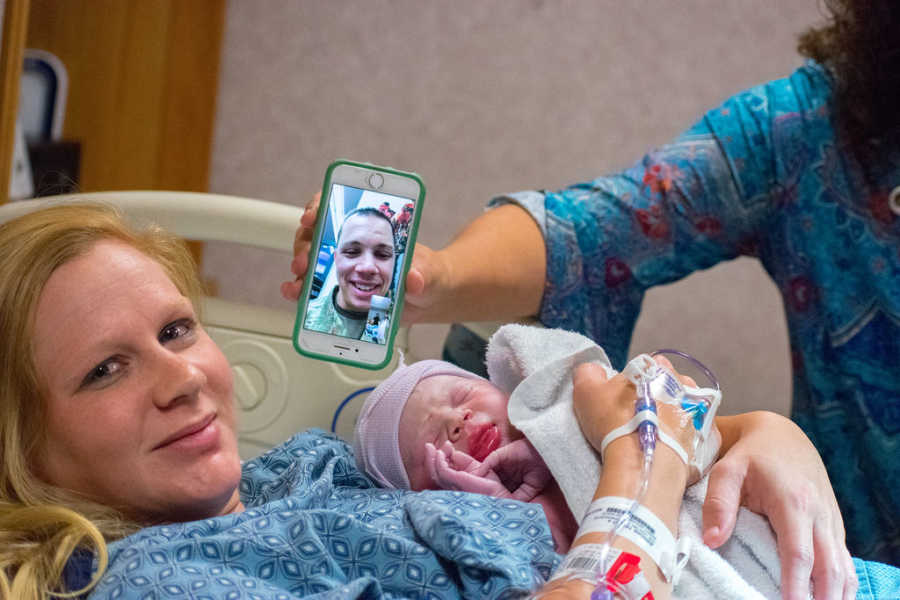 Woman smiles with newborn laying on her chest and iPhone held up next to her with military husband on facetime