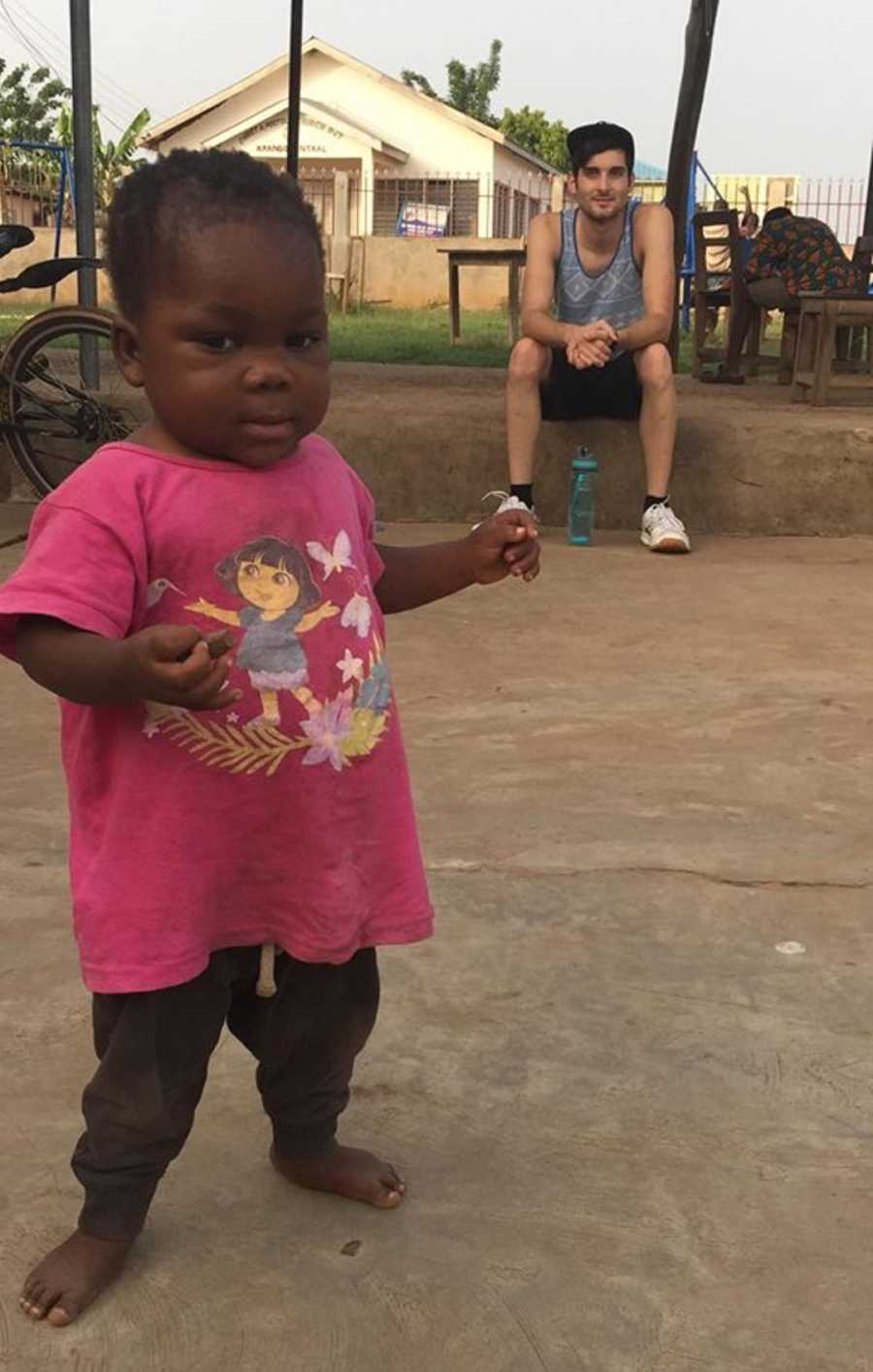 Adopted toddler from Africa stands barefoot while her father sits in background watching her