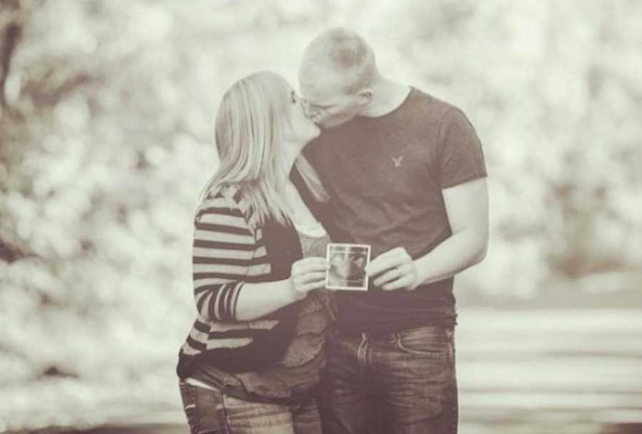 Husband and wife kiss as they hold ultrasound picture in front of them 