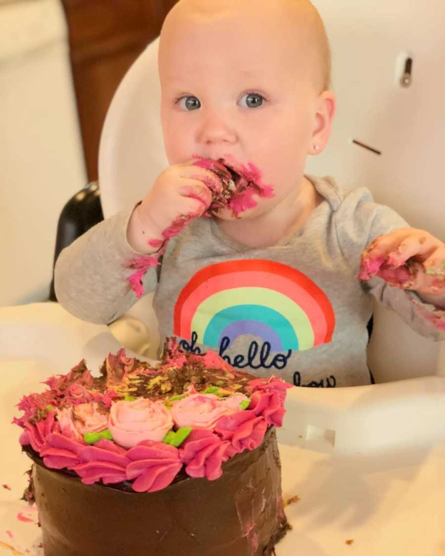 Baby sits in high chair eating cake with her hands with pink frosting all over her hands and face
