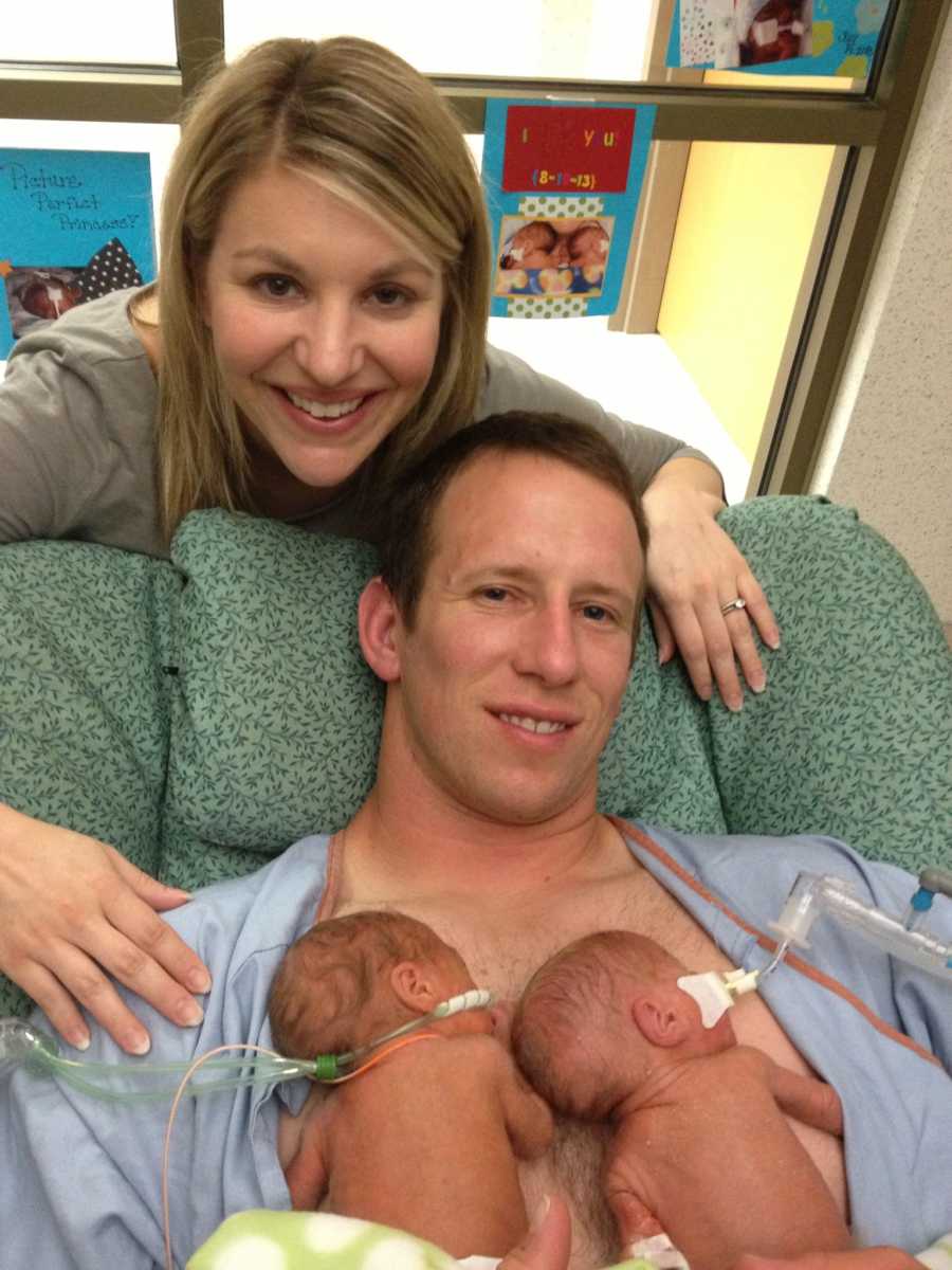 Wife smiles behind chair husband sits in with their newborns sleeping on his bare chest