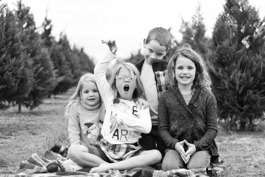 Four siblings sit on grass on blanket between Christmas trees