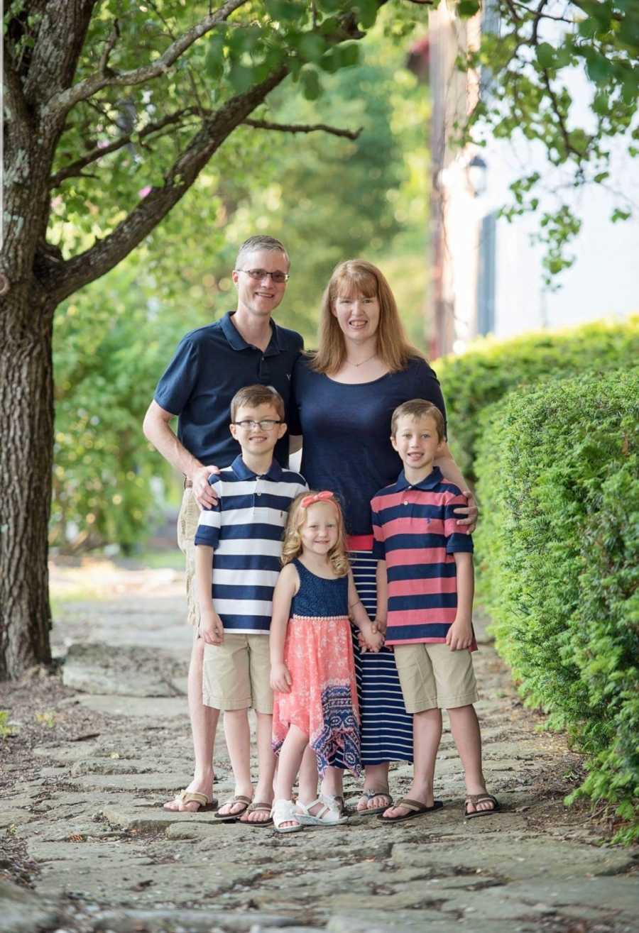 Husband and wife stand on sidewalk smiling with their two son's and daughter in front of them