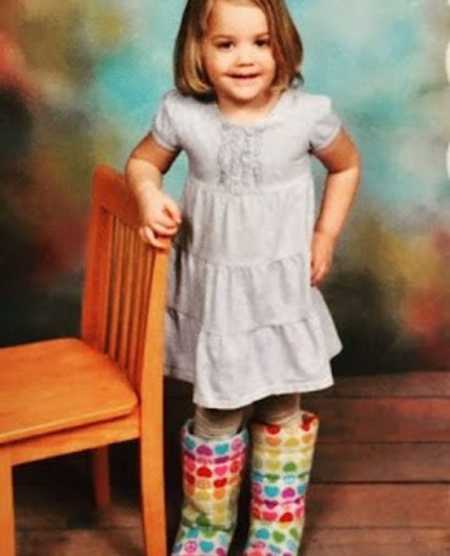 Little girl stands with arm on back rest of chair wearing dress and rainbow heart boots