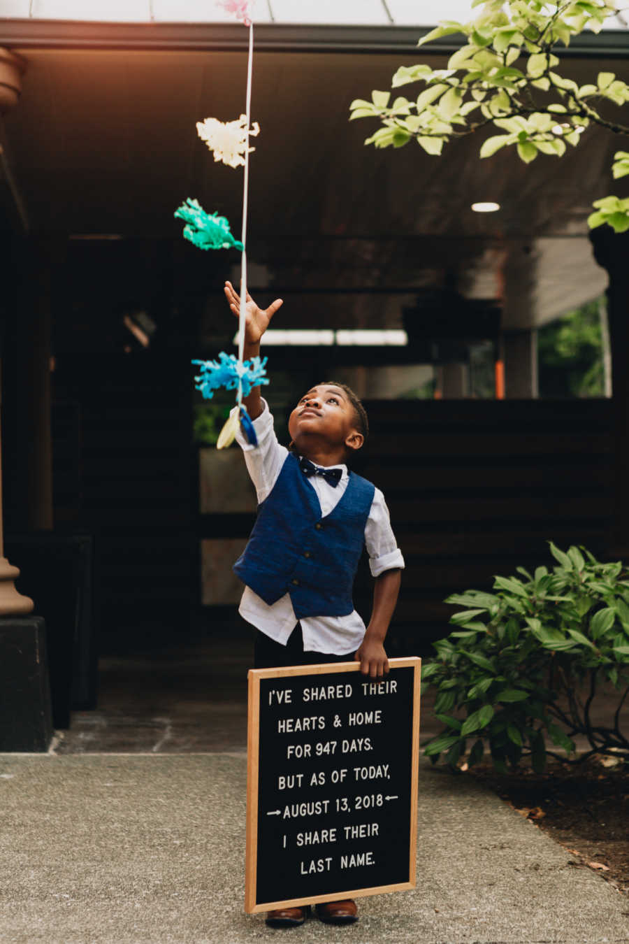 Little boy in blue vest and boy tie holds sign in one hand while reaching for balloon string