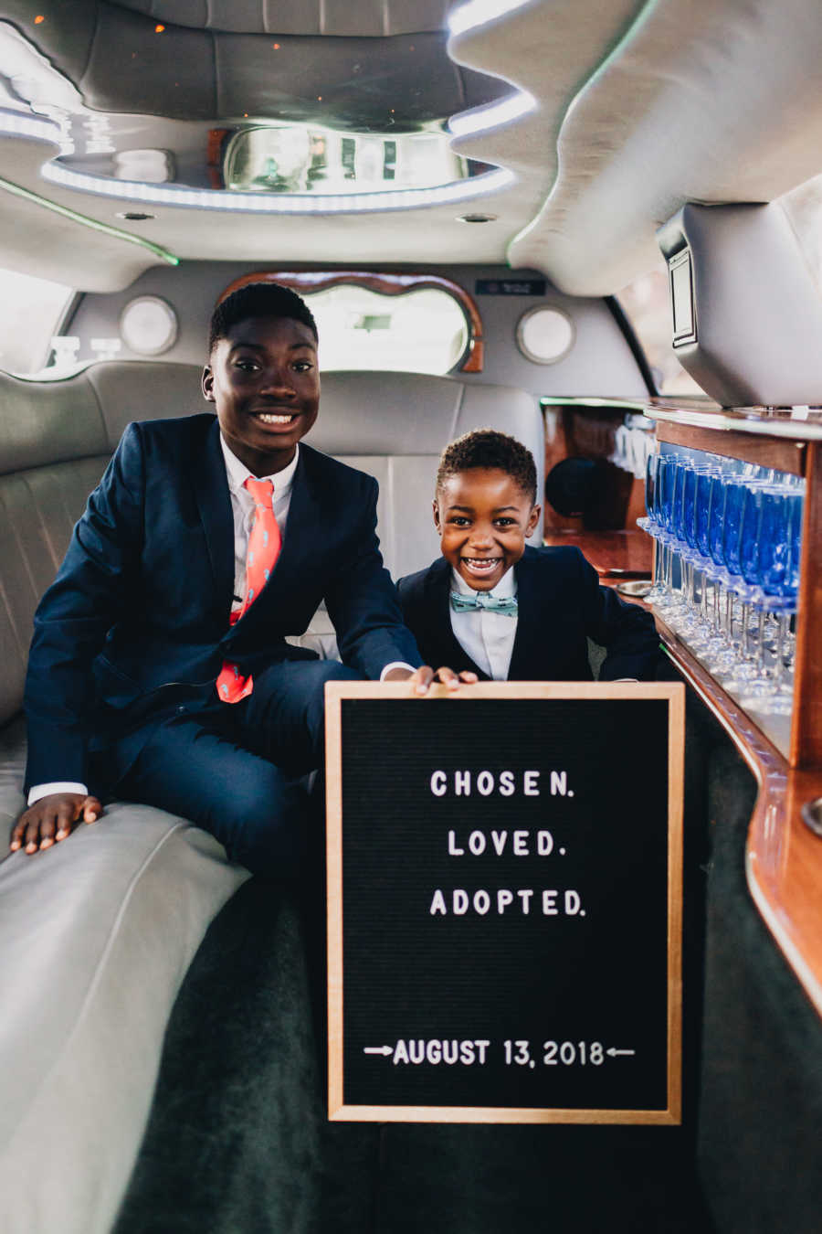 Biological brothers sit in limo with sign saying, "chosen, loved adopted" after being adopted by same parents