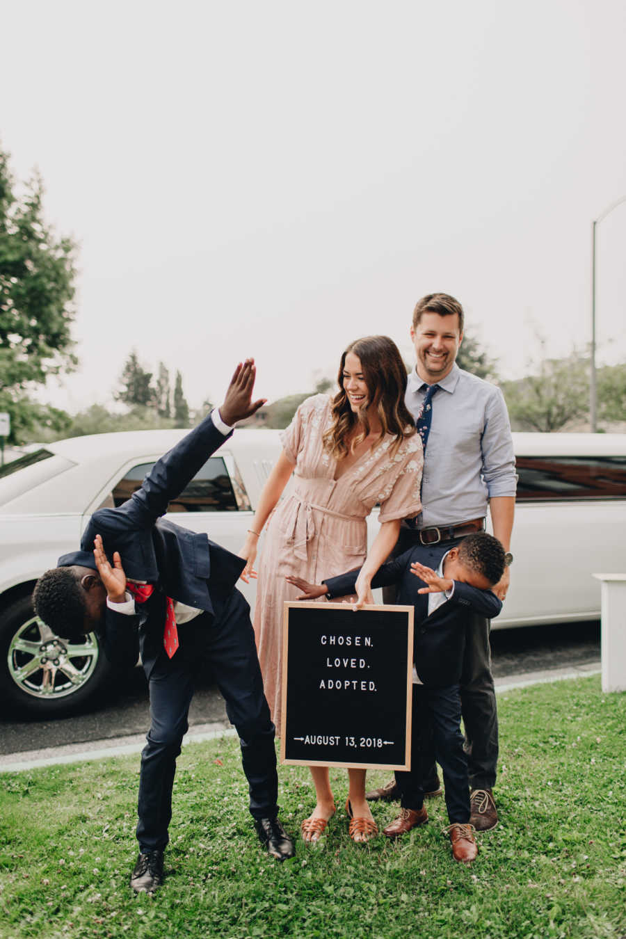 Husband and wife stand holding sign behind two adopted sons who are doing the dab pose