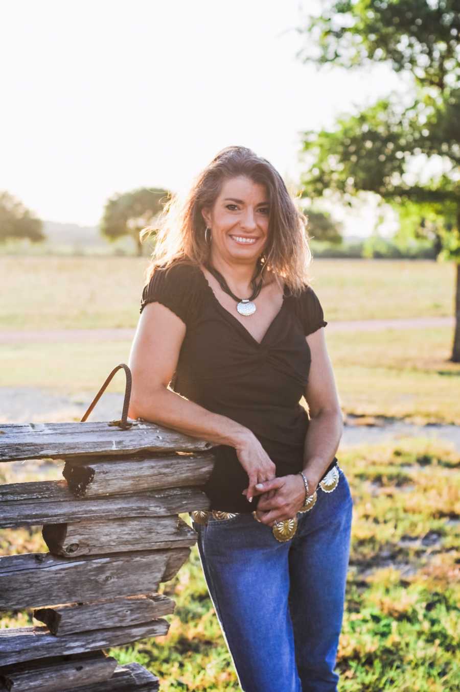 Woman who has abused drugs and been to jail stands smiling while leaning against wooden fence