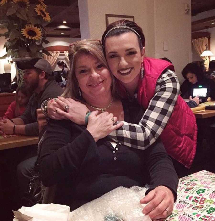 Daughter who found drugs in mother's purse stands hugging mother who is sitting at restaurant table