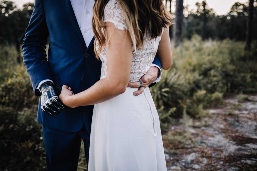 Bride and groom stand arm in arm while bride holds groom's bionic hand 