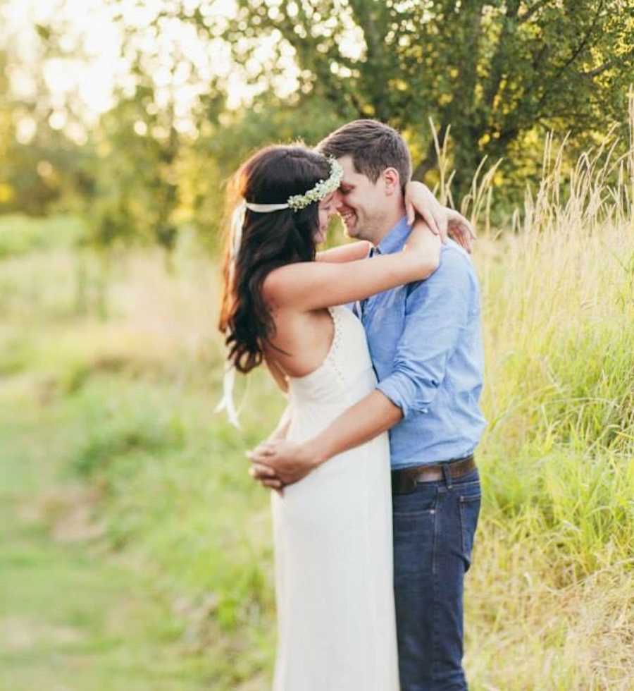 Bride and groom stand with arms wrapped around each other smiling in field