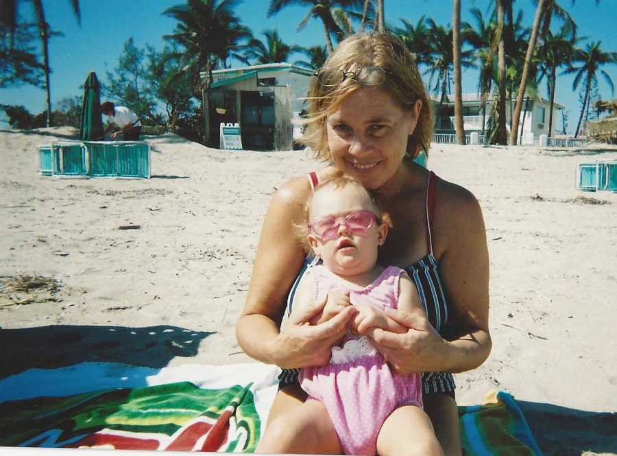 Woman sits kneeling on beach blanket with granddaughter on her lap