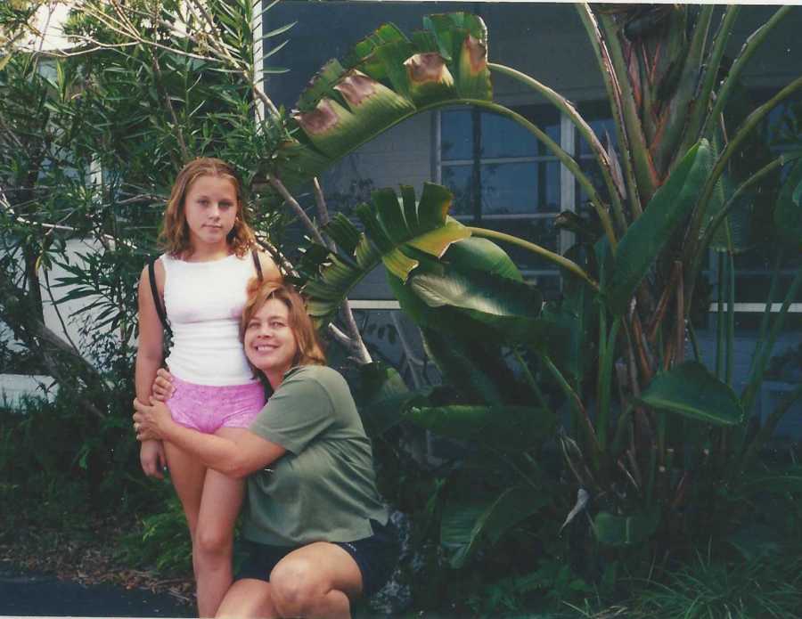 Teen standing in front of house while her mother crouches down and hugs her at her waist