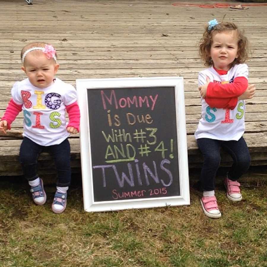 Two young sisters sit on porch with sign between them that says, "Mommy is due with #3 and #4 twins"