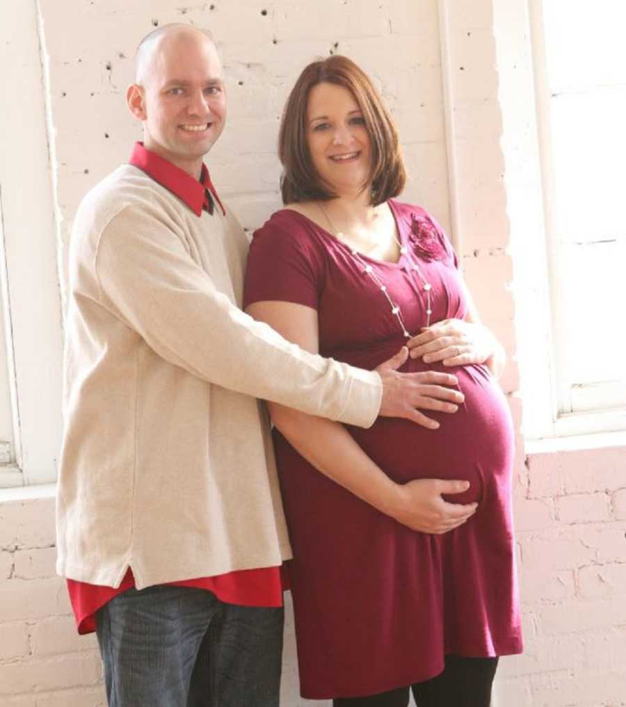 Pregnant woman stands smiling holding her stomach while husband stands beside her with hand on her stomach