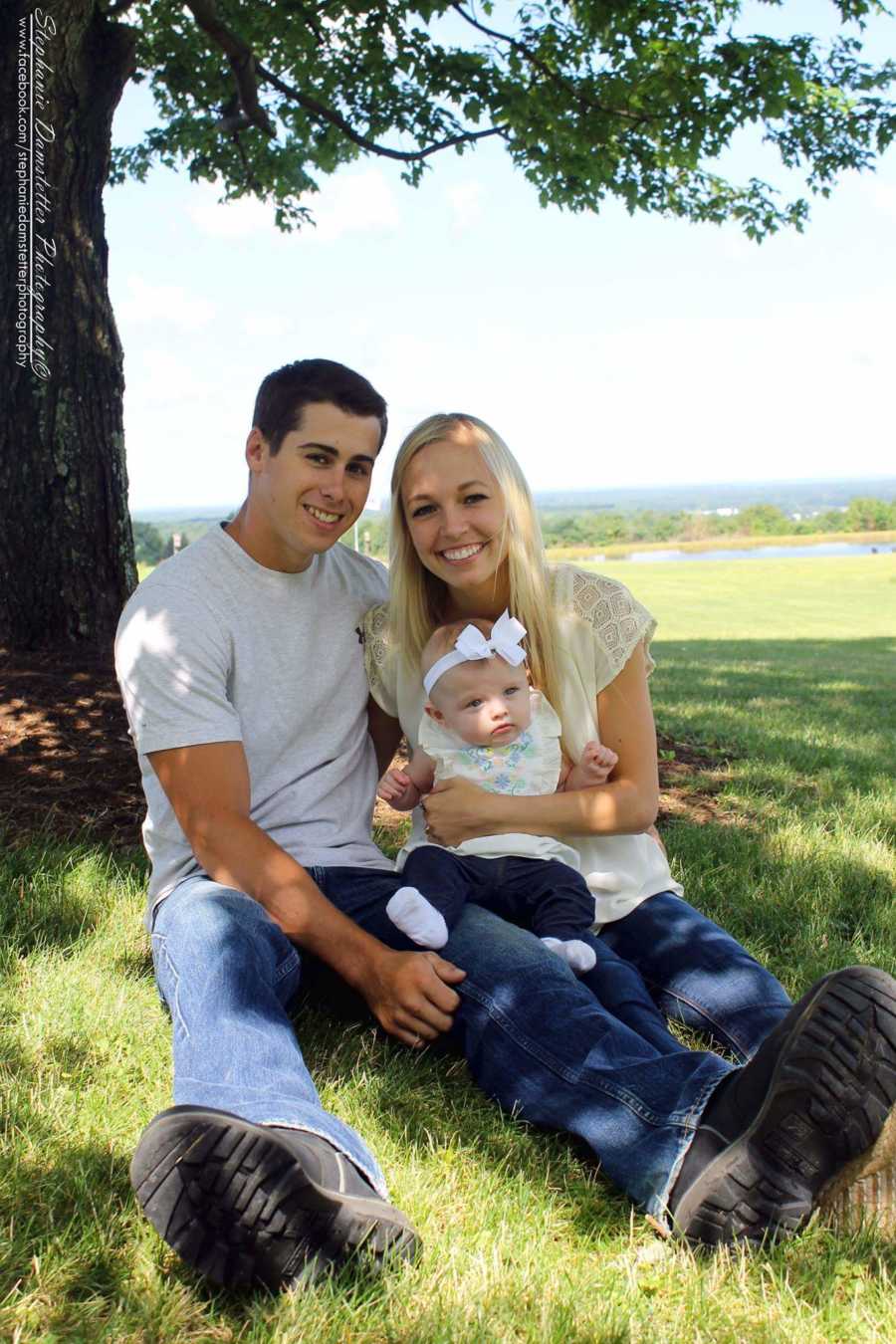 Husband and wife sit in grass with daughter in their lap