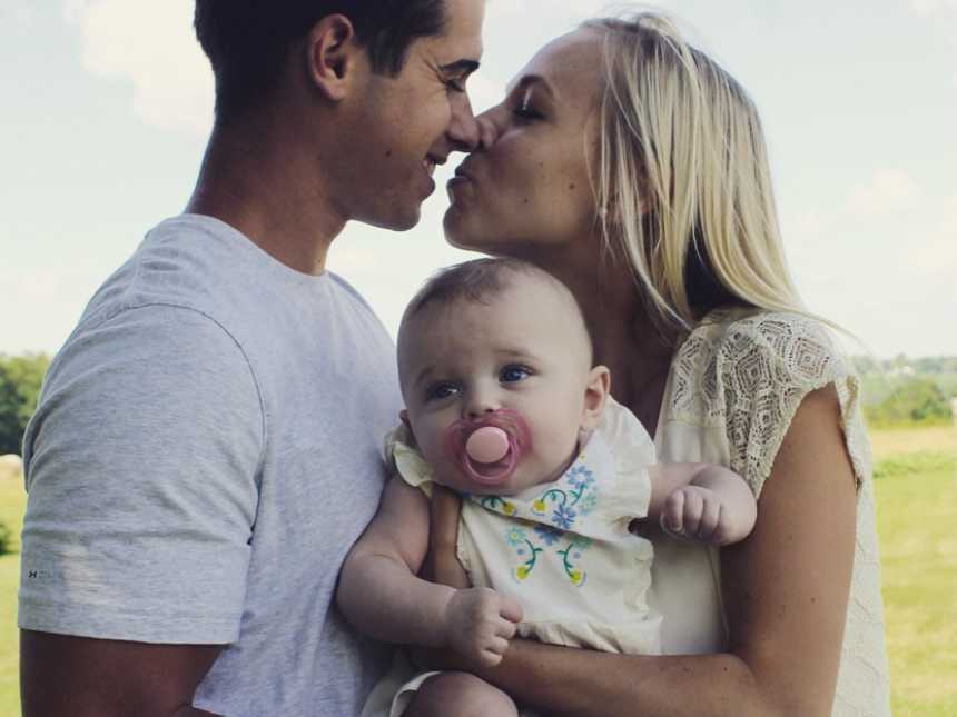 Husband and wife lean in to kiss and wife holds baby daughter
