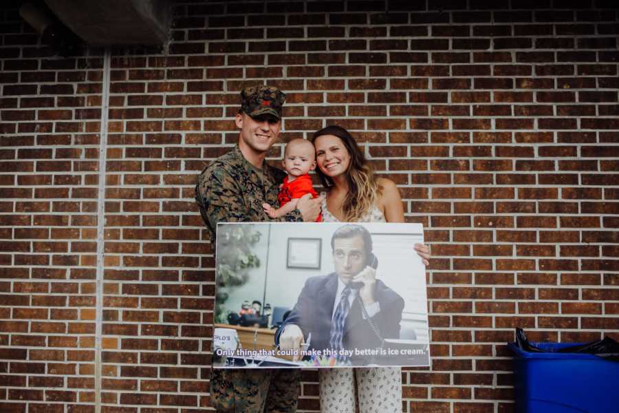 Soldier father holds son at return from deployment while wife holds sign from scene of The Office