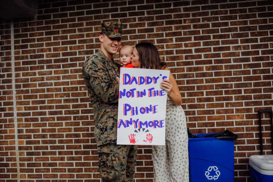 Army father holds infant son as wife kisses son's cheek and holds sign saying, "Daddy's not in the phone anymore"