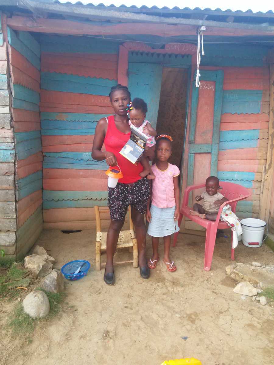 Poor woman in Haiti standing outside house with three children holding energy lamp man donated to her