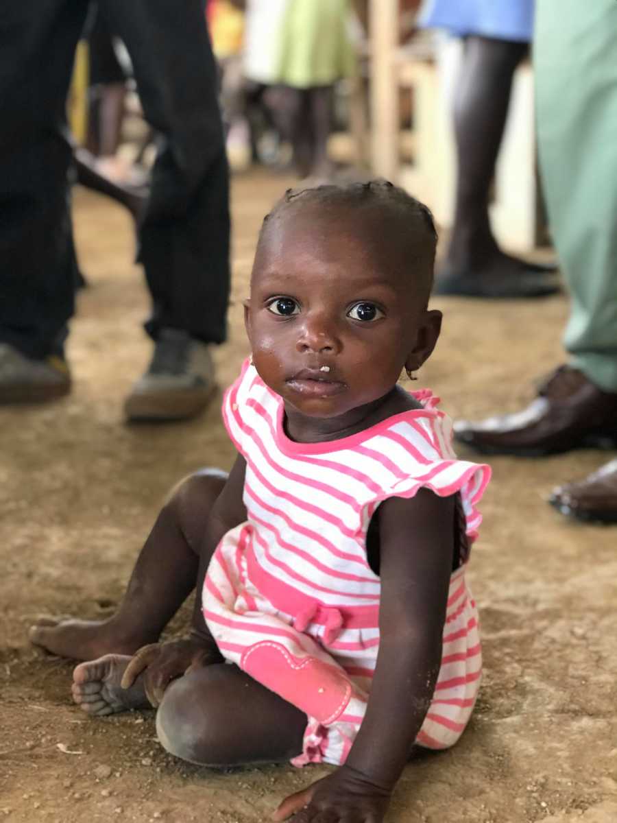 Infant Haitian orphan sitting on dirt ground in pink and white stripe dress