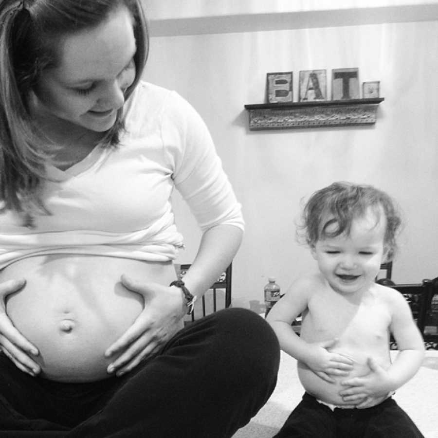 Pregnant mother sits on floor with hands on her stomach beside her toddler son who is copying her