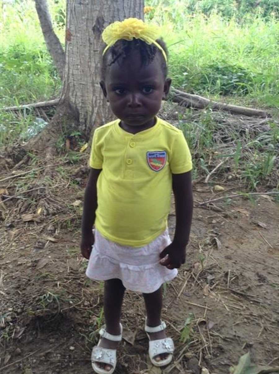 Haitian foster child stands near tree in bright yellow shirt in headband with white skirt and shoes
