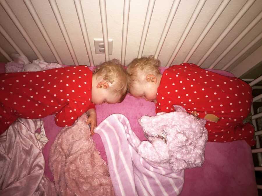 Twin girls lay head to head asleep in crib in red and white polka dot onesie