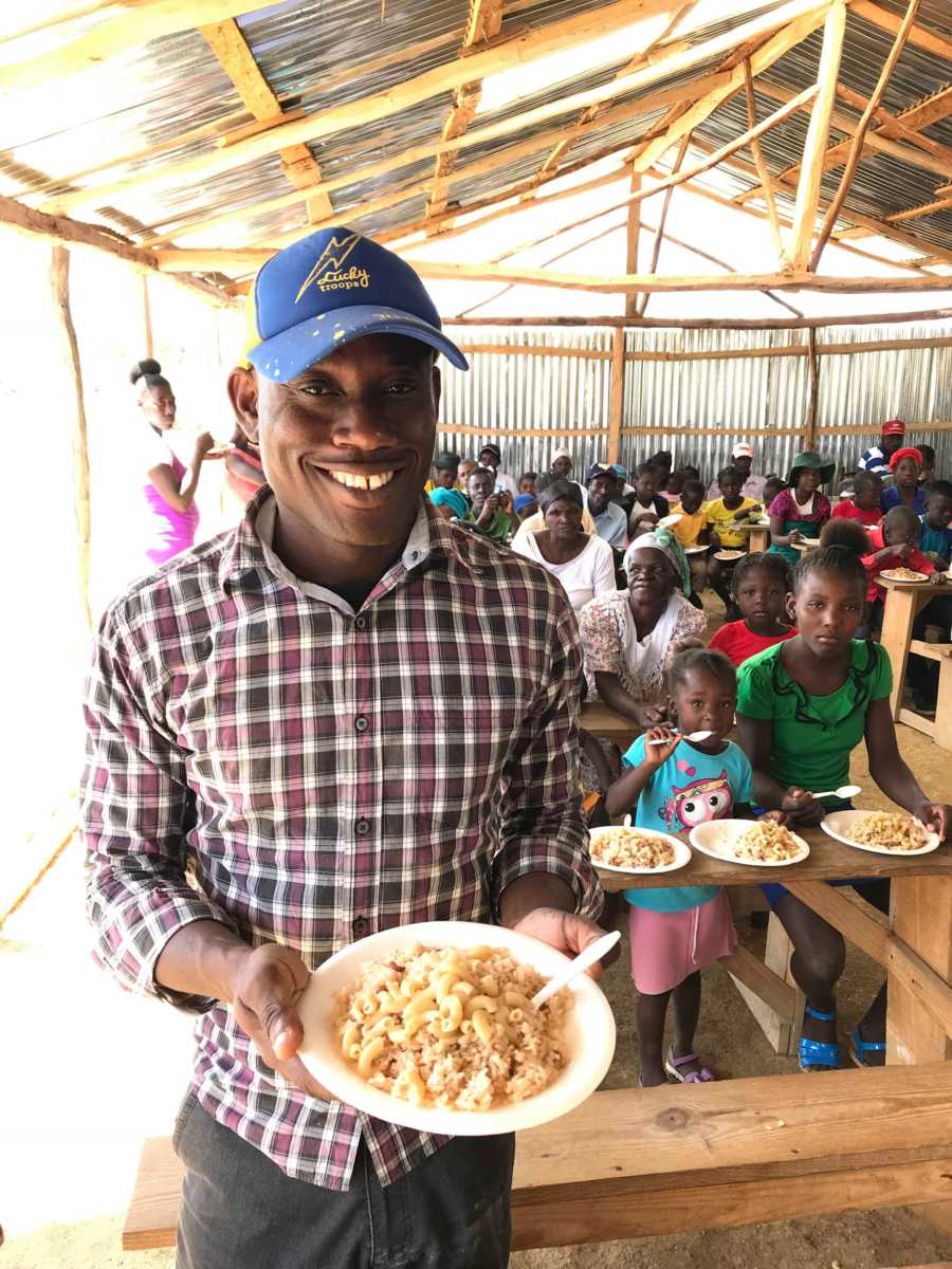 Man who fosters 62 children in Haiti stands holding bowl of food in front of orphans who are eating in outdoor cafeteria 