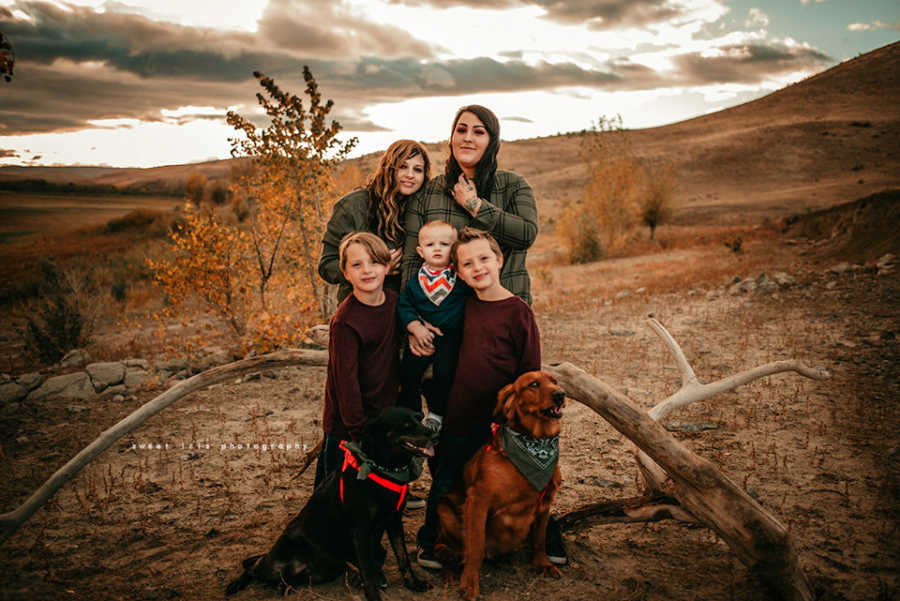 Wifes stand in desert with their baby son, one of the wife's twins and their two dogs