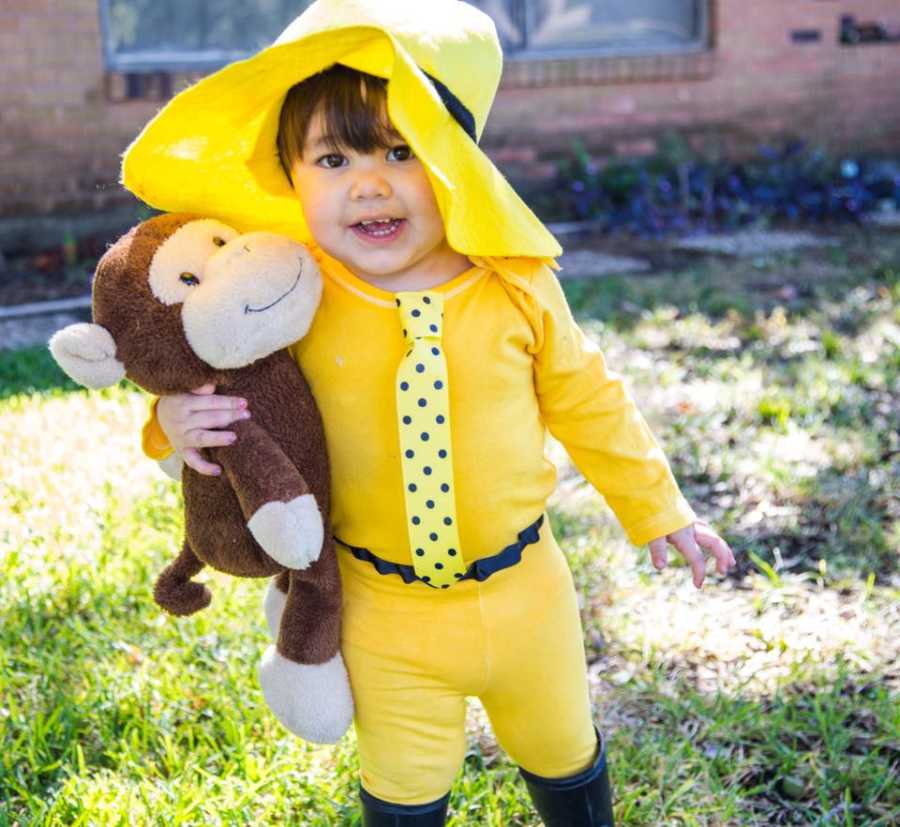 Toddler boy standing in yard dressed as the man in yellow hat holding onto stuffed monkey