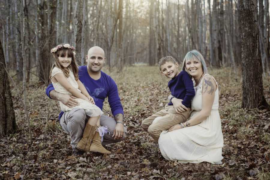 Husband and wife kneel in wooded area with their son and daughter sitting on their lap