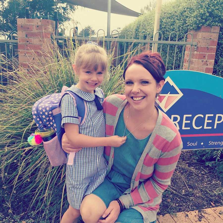 Young girl with backpack on stands smiling beside her mother outside of her rehab center