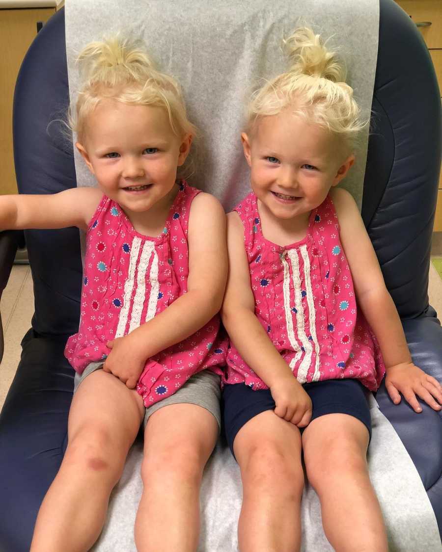 Twin girls sit in chair wearing matching pink tank tops 