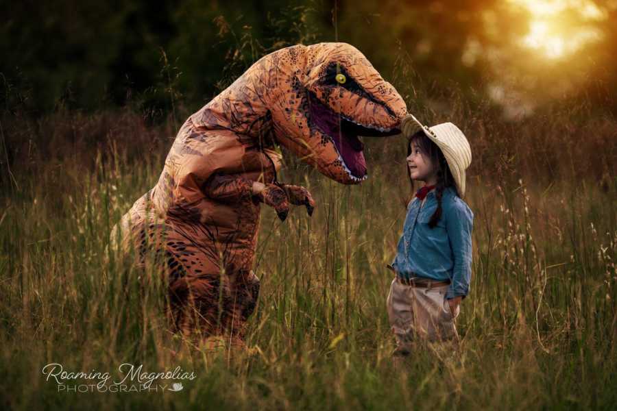 Young girl with autism stands in field in front of person in dinosaur costume