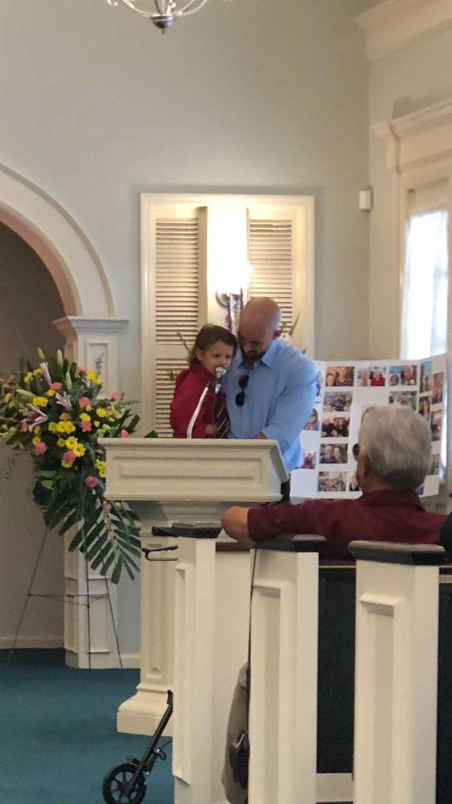 Father stands holding son at podium at his wife and daughter's funeral