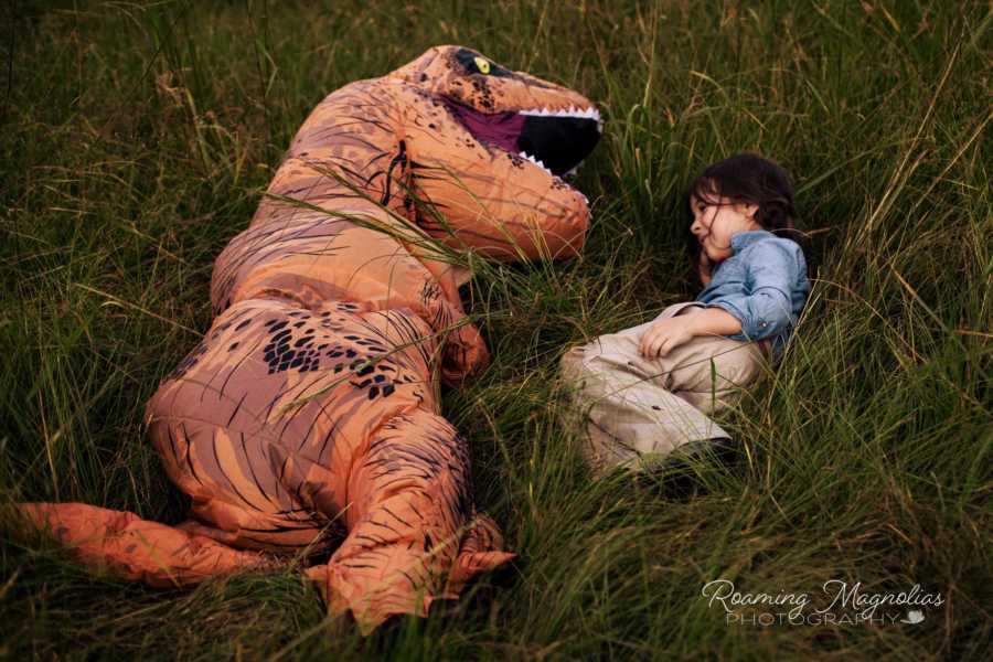 Young girl with autism lies in field beside person in dinosaur costume