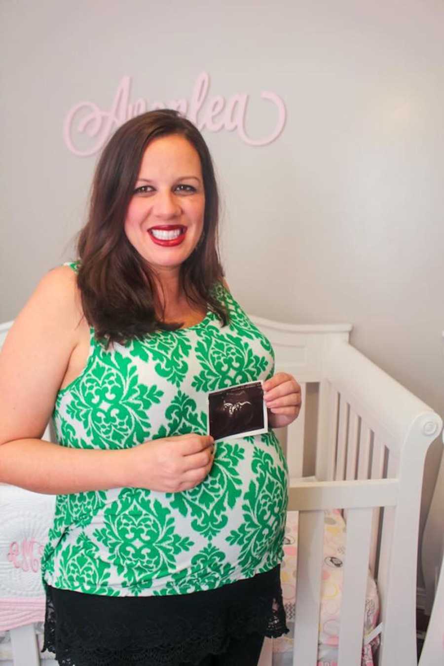 Pregnant woman standing in front of crib holding ultrasound picture over her stomach