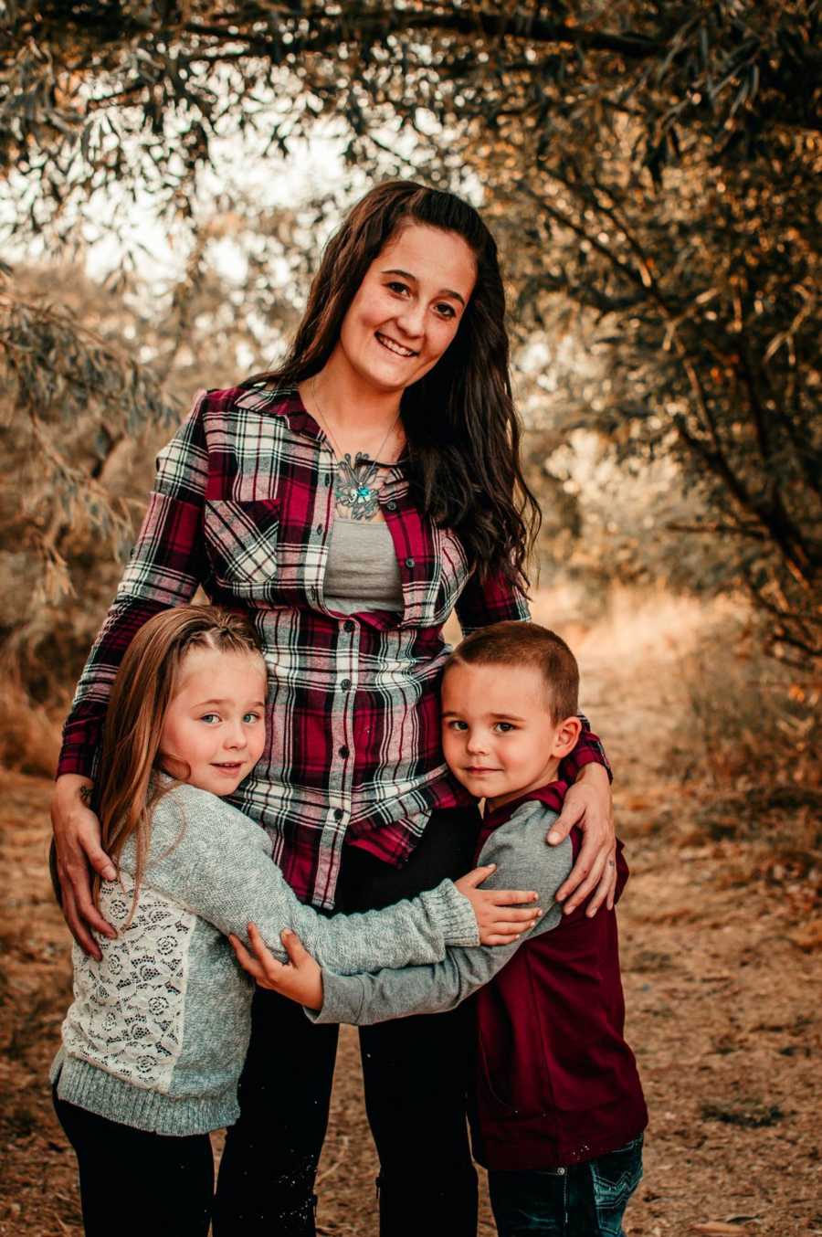 Woman stands smiling with her two children at her side hugging her waist