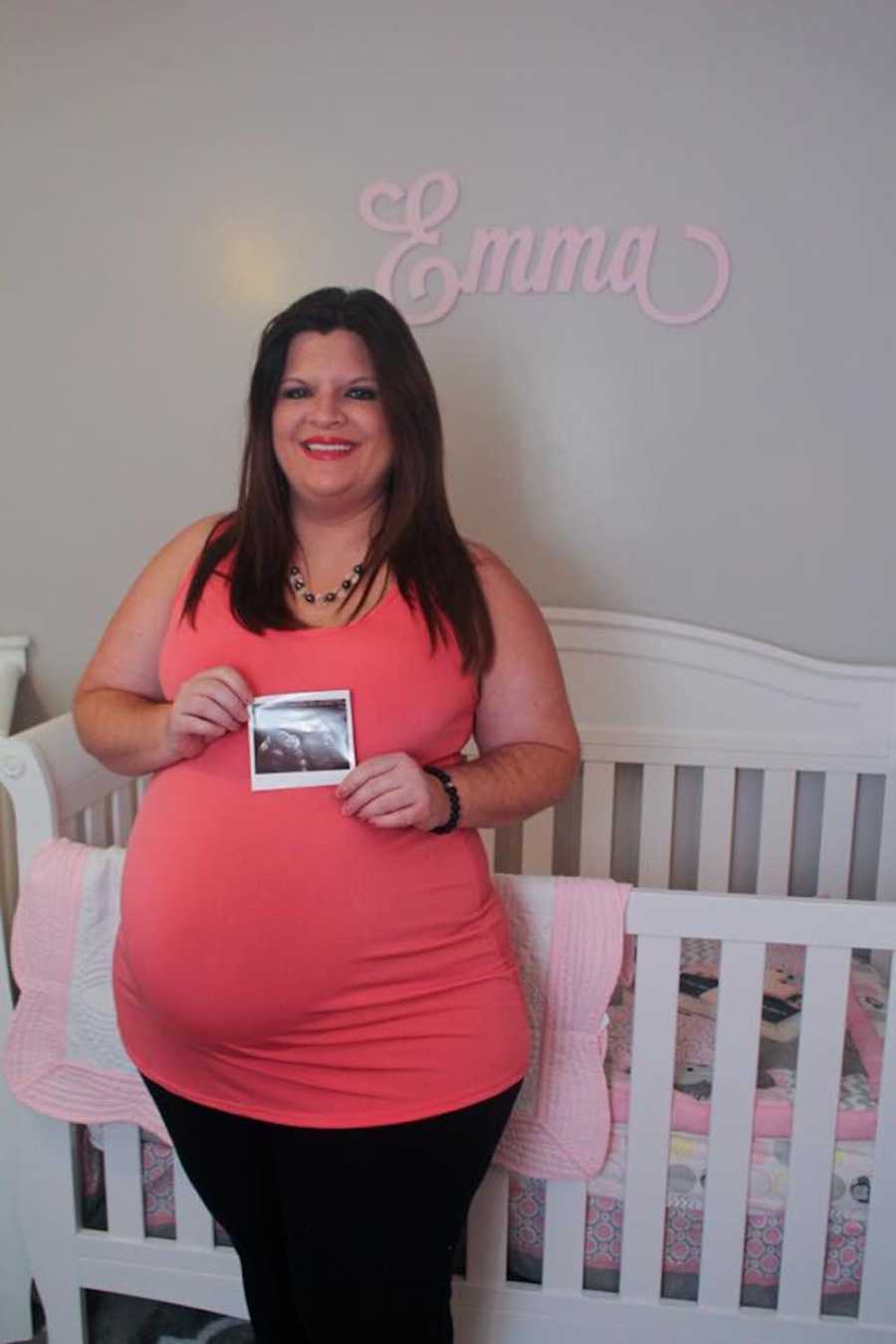 Pregnant woman smiling in front of crib holding ultrasound picture in front of her