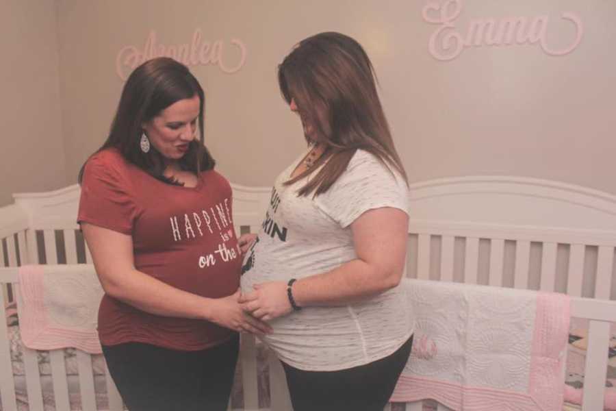 Pregnant wives stand stomach to stomach in front of cribs