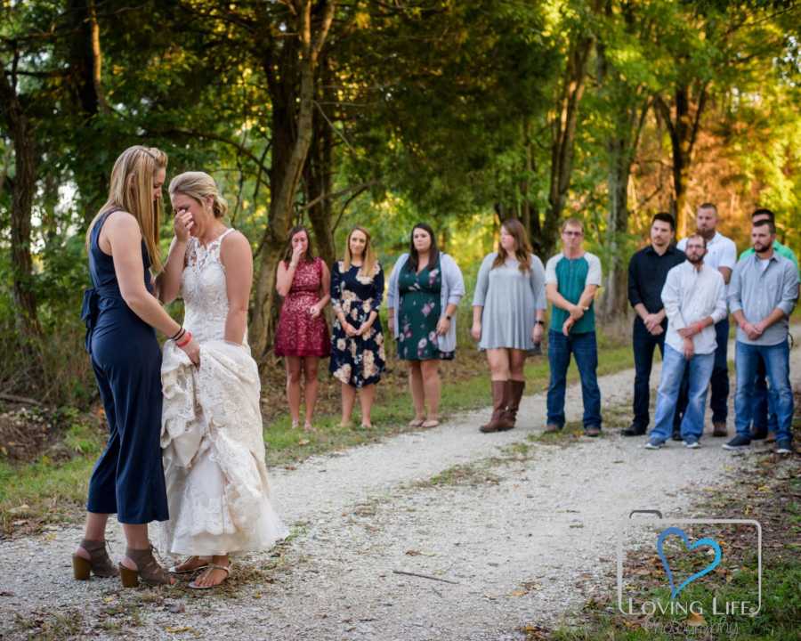 Woman in wedding gown cries while sister holds her gown and family members watch h