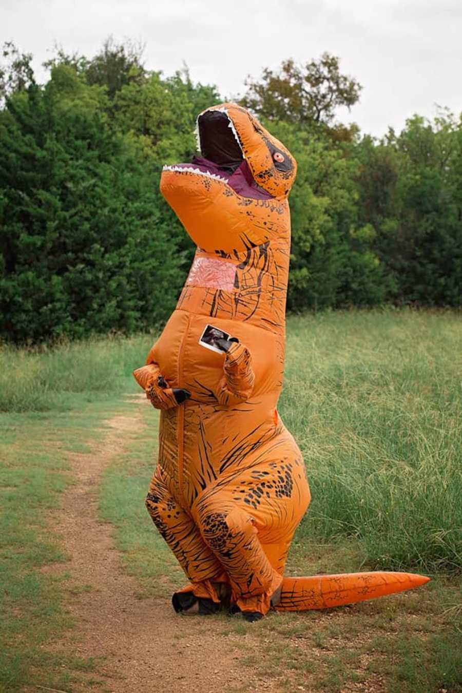 Person in dinosaur costume stands in field holding ultrasound picture