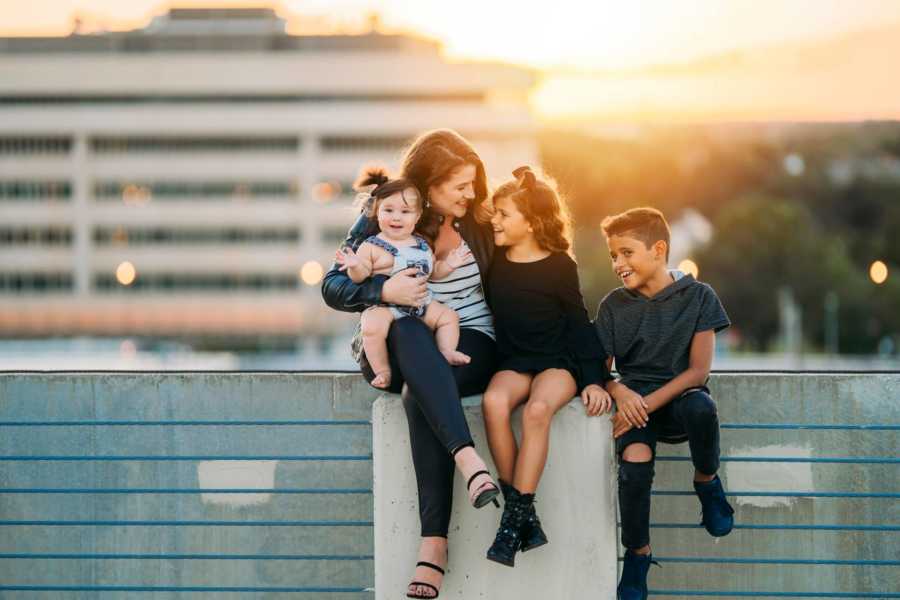 Woman sits on post of parking garage beside her children and her boyfriend's daughter