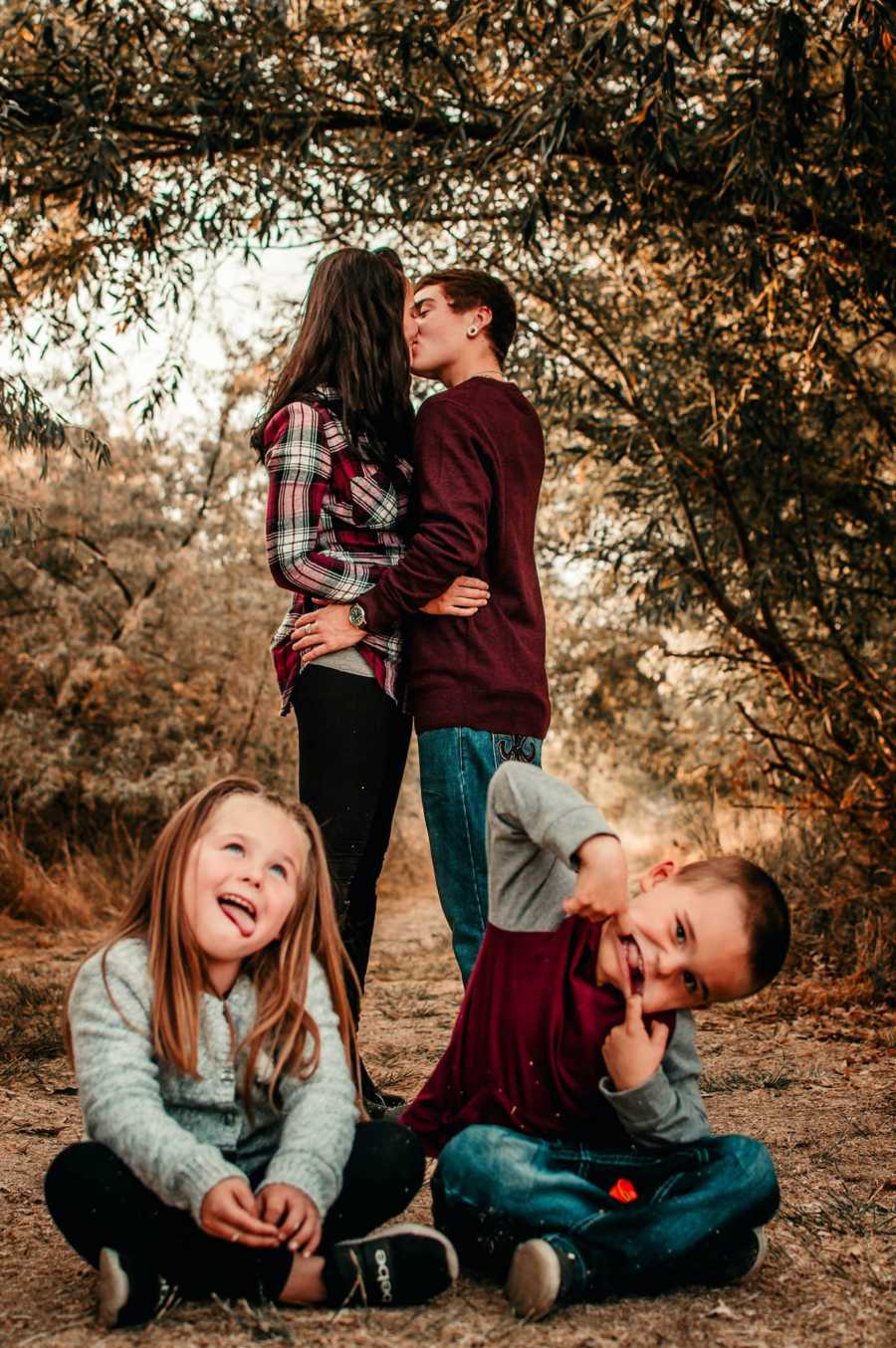 Girlfriend and transgender boyfriend kiss while girlfriend's kids sit on ground in front of them making funny faces