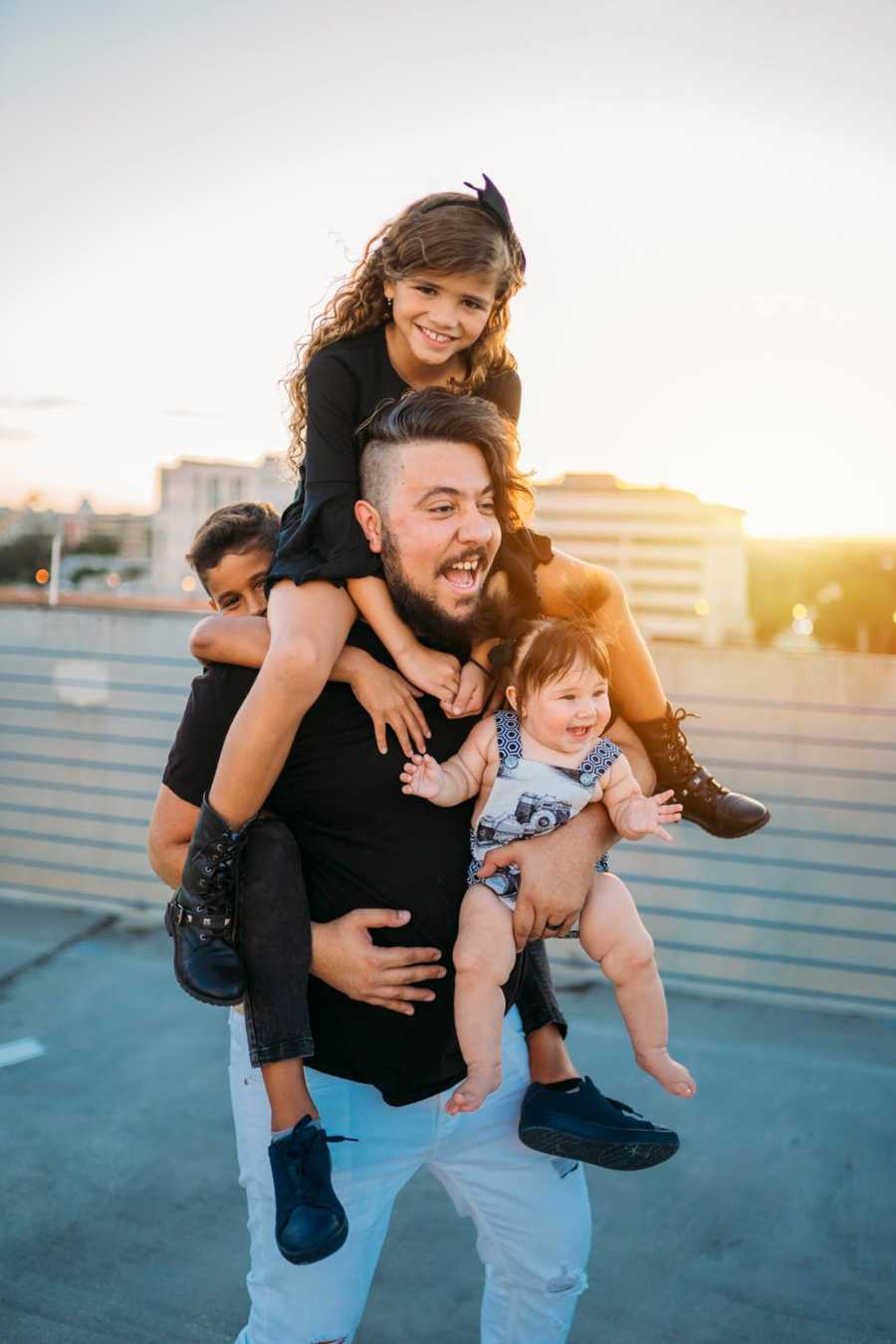 Man stands holding his baby daughter along with girlfriend's daughter on his shoulders and son on his back